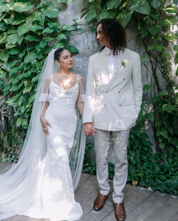 Vanessa Hudgens in a minimalist cowl-neck satin mermaid wedding gown with a cathedral veil standing with her groom against a lush green ivy backdrop