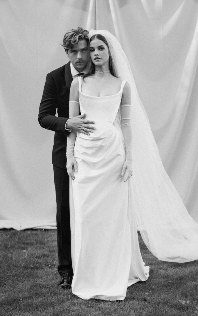 Barbara Palvin in her Vivienne Westwood minimalist square-neck column wedding gown with sheer long gloves and cathedral veil, in a black and white portrait with her groom