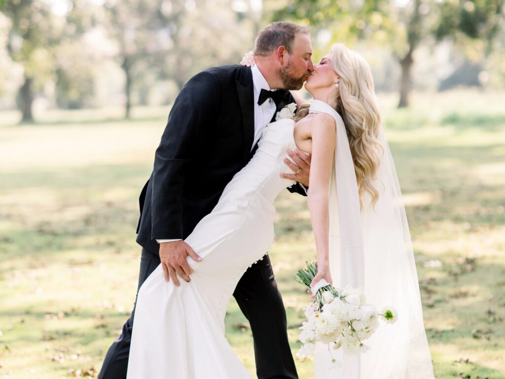 Groom dipping and kissing bride Ashley VanZant outdoors, her white column gown and cathedral veil flowing as she holds a white bouquet of peonies and daisies