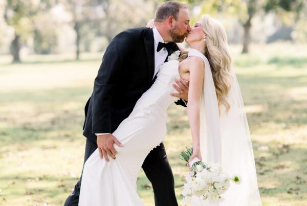 Groom dipping and kissing bride Ashley VanZant outdoors, her white column gown and cathedral veil flowing as she holds a white bouquet of peonies and daisies