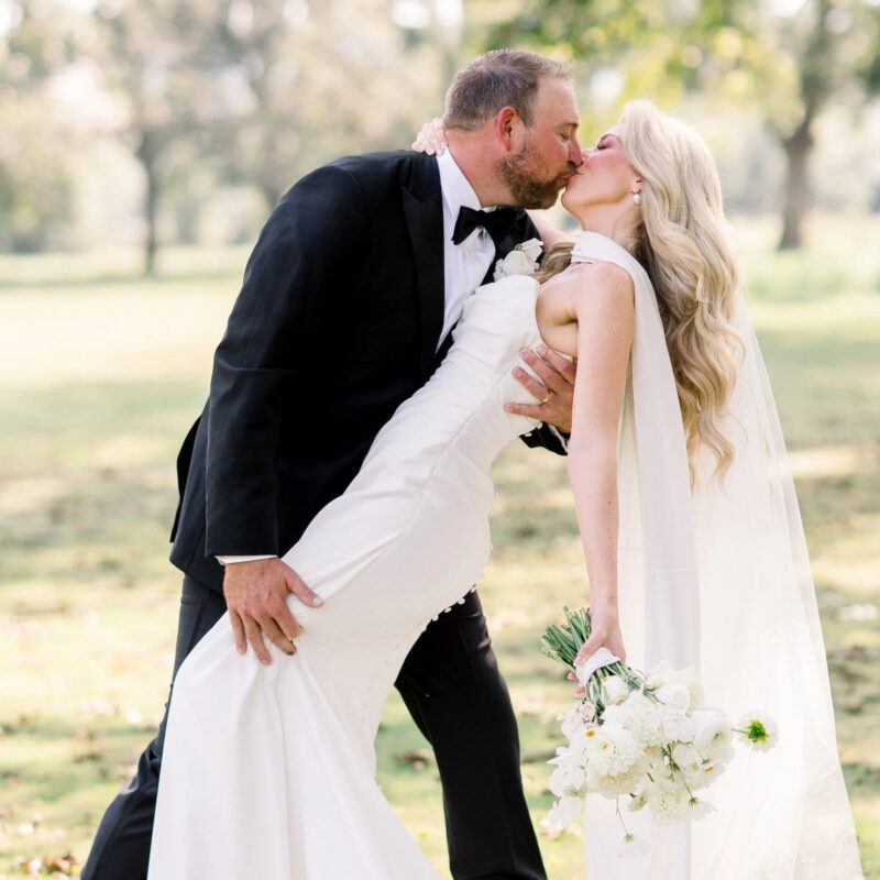 Groom dipping and kissing bride Ashley VanZant outdoors, her white column gown and cathedral veil flowing as she holds a white bouquet of peonies and daisies