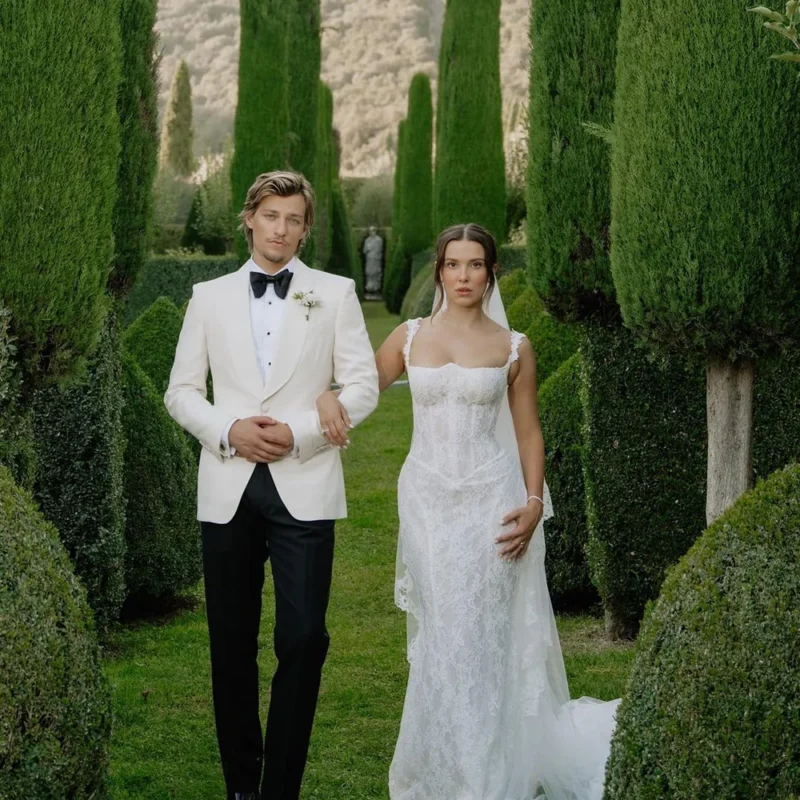 Millie Bobby Brown and Jake Bongiovi on their wedding day, with Millie wearing a fitted Galia Lahav lace gown with lace-trimmed straps and a cathedral veil, surrounded by tall cypress trees