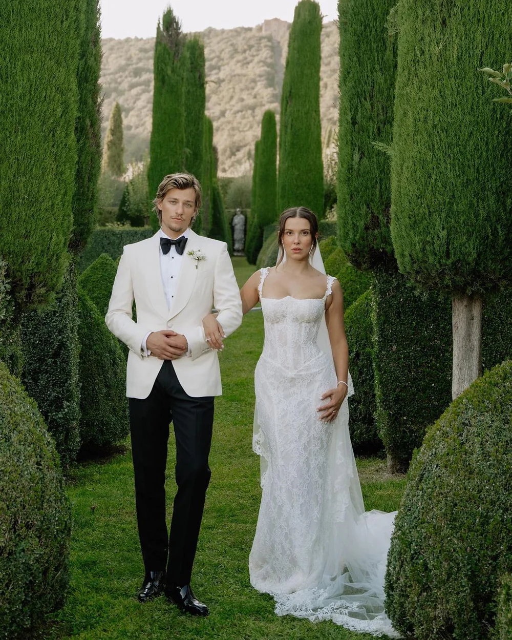 Millie Bobby Brown and Jake Bongiovi on their wedding day, with Millie wearing a fitted Galia Lahav lace gown with lace-trimmed straps and a cathedral veil, surrounded by tall cypress trees