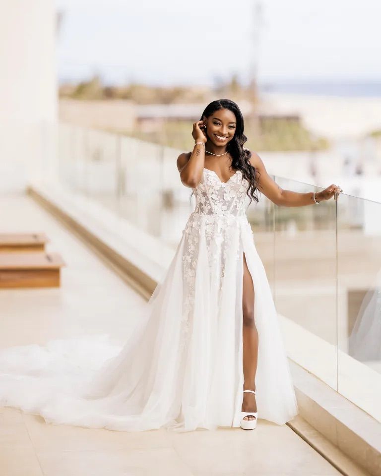 Simone Biles in her Galia Lahav sweetheart lace appliqué wedding gown with a thigh-high slit and cathedral train on a sunny outdoor terrace