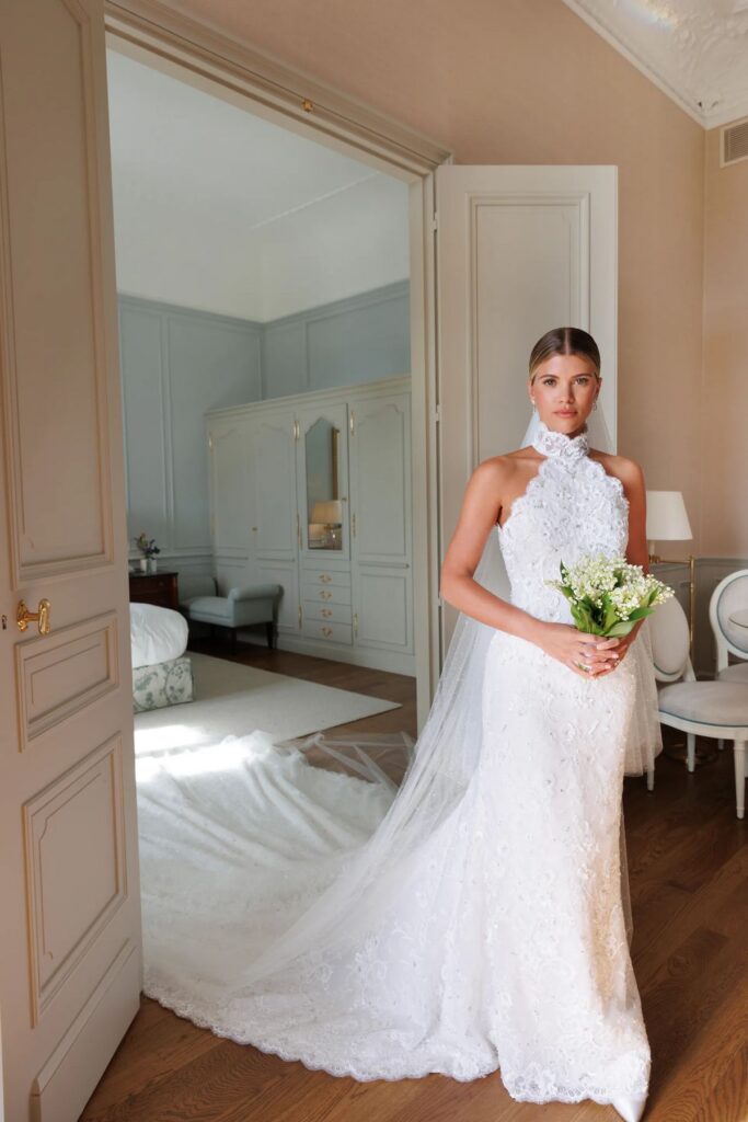 Sofia Richie in her custom Chanel wedding gown, a halter-neck lace fit-and-flare dress with a long train, holding a lily of the valley bouquet