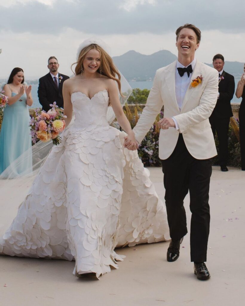 Joey King and Steven Piet celebrating after their ceremony, with Joey wearing an Oscar de la Renta strapless sweetheart ball gown covered in sculptural petal appliqués, holding a colorful bouquet overlooking the water
