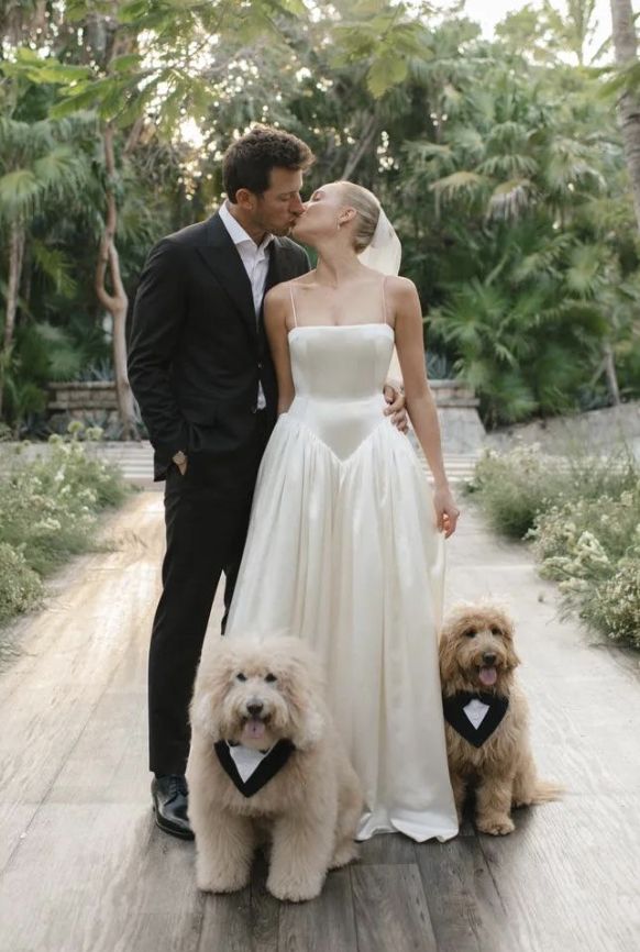 Alex Cooper in a sleek spaghetti-strap square-neck satin ball gown with a structured bodice, kissing her groom outdoors with their two goldendoodles wearing tuxedo bandanas