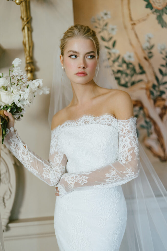 Bride wearing an off-the-shoulder lace wedding gown with sheer long sleeves and a fitted silhouette, holding a white bouquet at La Mariée Boutique Tulsa