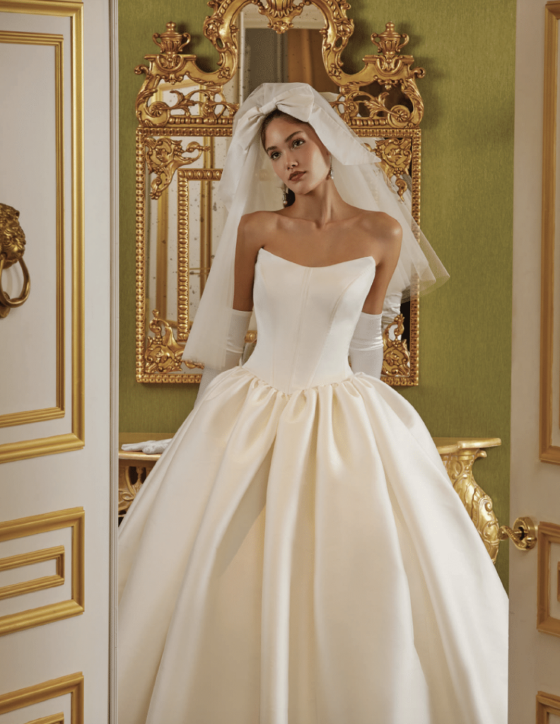 Strapless structured satin ball gown wedding dress with a bow-tied veil and short bridal gloves in an ornate gilded interior at La Mariée Boutique Tulsa