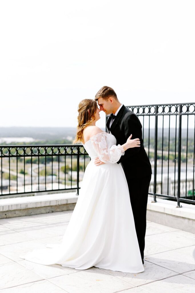 Bride in off-shoulder lace wedding gown and groom in black tuxedo sharing intimate moment on rooftop terrace overlooking Tulsa