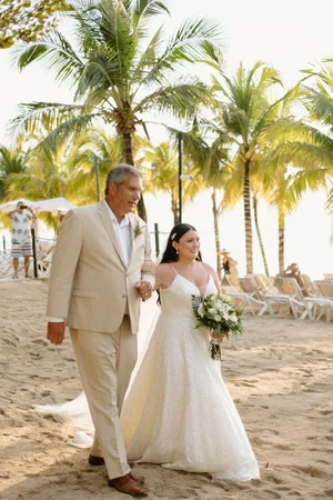 Bride walks down a sandy beach aisle on the arm of her father beneath swaying palm trees, wearing a lace A-line gown and carrying a white and greenery bouquet
