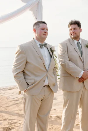 Groom in a tan suit and sage bow tie stands at the altar with a groomsman, watching for the bride's arrival at a beach ceremony