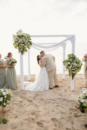 Bride and groom share their first kiss at the altar, framed by a white draped arch with white and green tropical florals, bridesmaids in sage dresses visible on either side