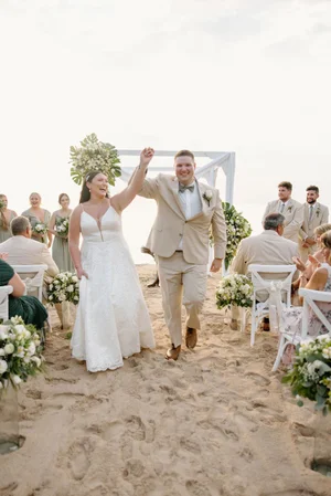 Bride and groom walk back down the aisle after being pronounced married, hands raised in celebration, guests cheering on either side at their Cozumel beach ceremony