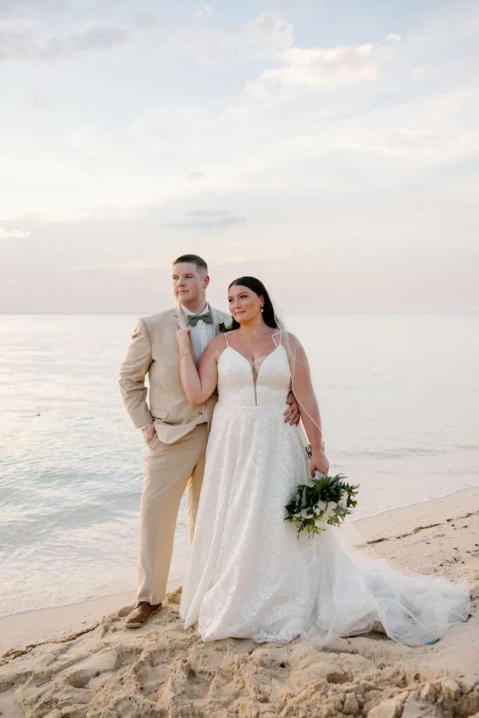 Bride and groom stand together on a sandy beach at golden hour, bride in a lace A-line gown with a deep V-neckline holding a white and greenery bouquet, groom in a tan suit with bow tie