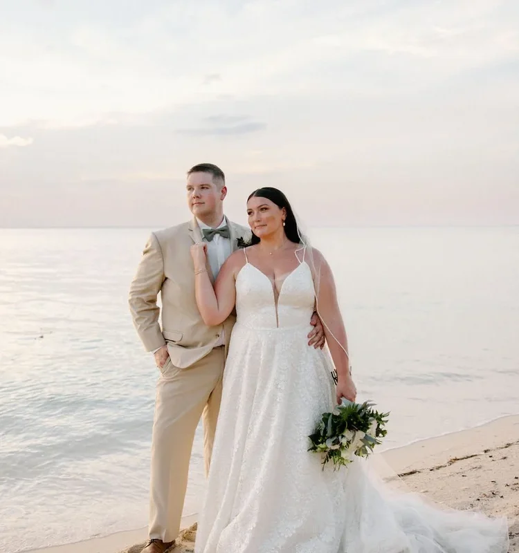 Bride and groom stand together on a sandy beach at golden hour, bride in a lace A-line gown with a deep V-neckline holding a white and greenery bouquet, groom in a tan suit with bow tie