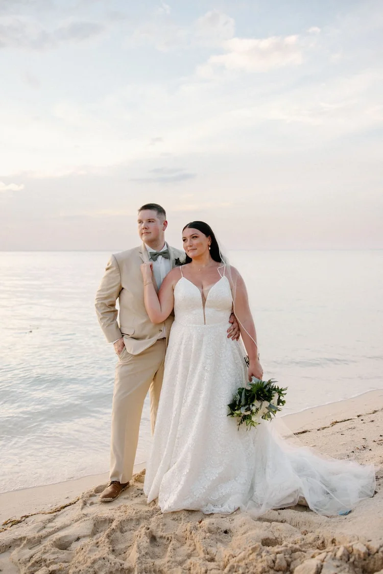 Bride and groom stand together on a sandy beach at golden hour, bride in a lace A-line gown with a deep V-neckline holding a white and greenery bouquet, groom in a tan suit with bow tie