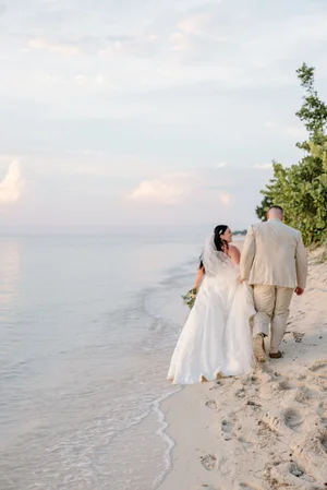 Bride and groom walk hand in hand along the shoreline at sunset, bride carrying her gown and bouquet as waves wash the sand behind them