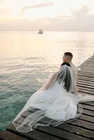 Bride sits alone at the end of a wooden dock overlooking the calm Caribbean Sea at sunset, cathedral veil spread across the weathered boards, a sailboat visible in the distance