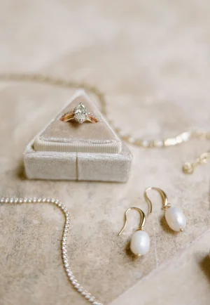 Flat lay detail shot of the bride's rose gold oval engagement ring in a velvet box, pearl drop earrings, and a delicate diamond tennis necklace