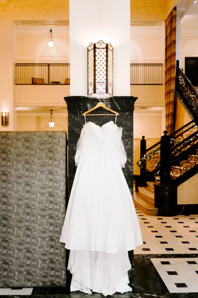 Off-shoulder a-line wedding dress with sheer floral lace sleeves hanging in ornate hotel lobby, La Mariée Tulsa bridal boutique