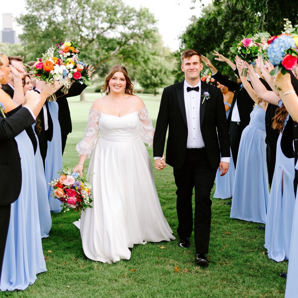Bride and groom walking through floral arch created by wedding party holding colorful bouquets at Tulsa Country Club