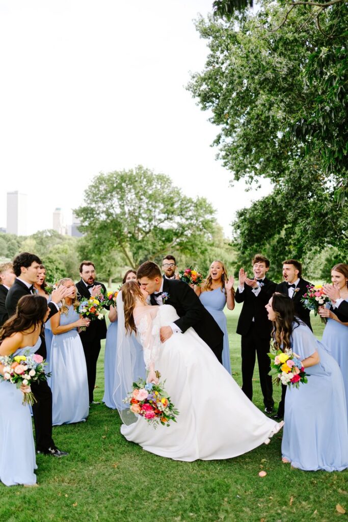 Groom dipping bride for a kiss surrounded by cheering wedding party in periwinkle bridesmaid dresses and black tuxedos