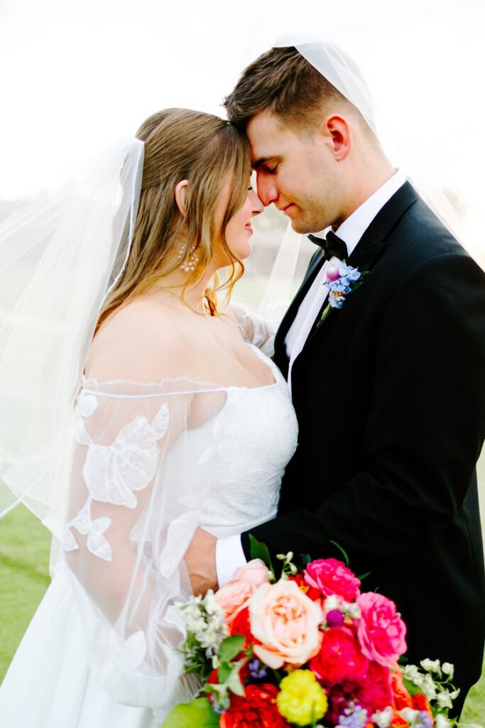 Bride and groom touching foreheads under veil with colorful bridal bouquet, intimate wedding portrait