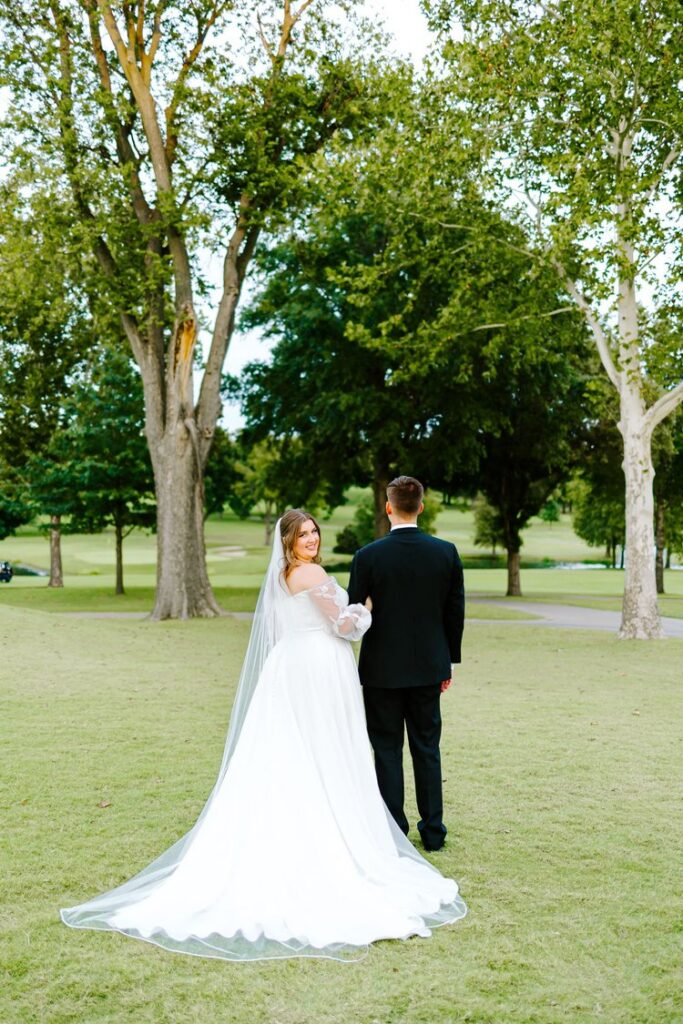 Bride in a-line wedding gown with cathedral veil walking hand-in-hand with groom through Tulsa Country Club golf course