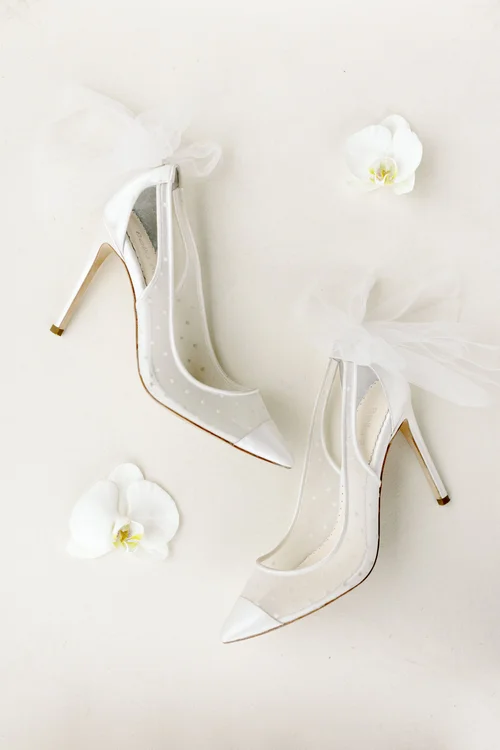 Bella Belle bridal heels with sheer polka dot mesh panels and oversized tulle bow detail, styled with white orchid blooms on a cream background