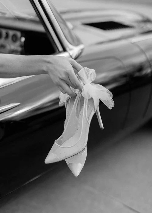 Black and white close-up of a bride's hand holding Bella Belle pointed-toe heels with tulle bow out the window of a vintage black convertible
