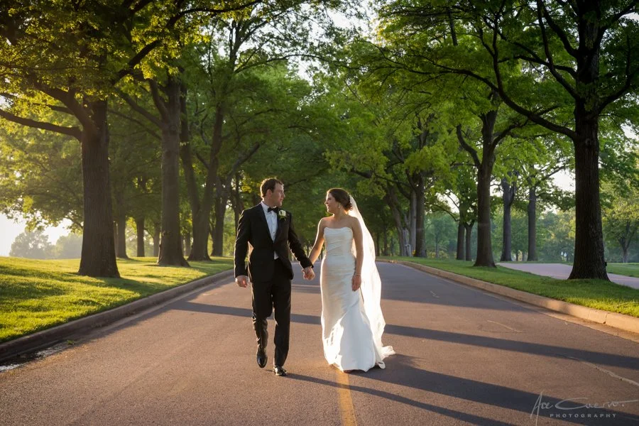 Bride and groom walking hand in hand down a tree-lined boulevard at golden hour after their Tulsa wedding, bride in a strapless satin mermaid gown with cathedral veil, groom in a black tuxedo