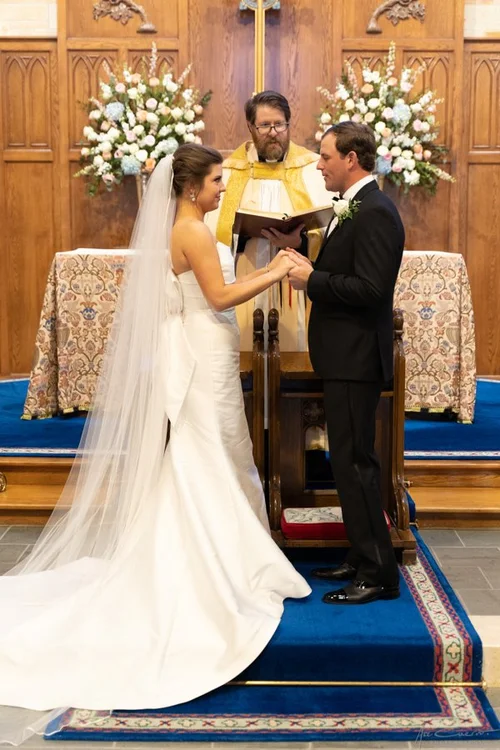 Bride and groom exchanging vows at the altar of St. John's Episcopal Church in Tulsa, bride in a strapless satin gown with cathedral veil, officiant holding ceremony book behind them flanked by pastel floral arrangements