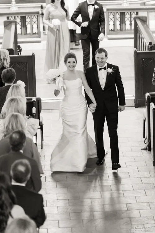Black and white photo of bride and groom walking back down the aisle at St. John's Episcopal Church in Tulsa, bride in a strapless satin mermaid gown beaming with joy, guests seated in wooden pews on either side