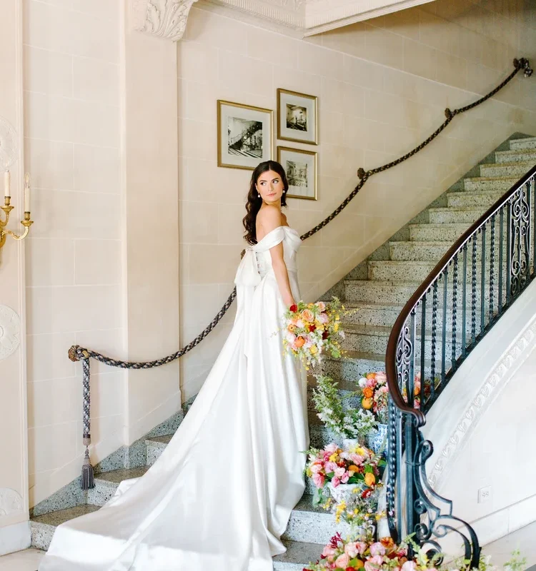 Bride in an off-the-shoulder satin A-line wedding gown with cathedral train standing on a grand staircase, looking back over her shoulder, surrounded by lush colorful floral arrangements in blue and white vases