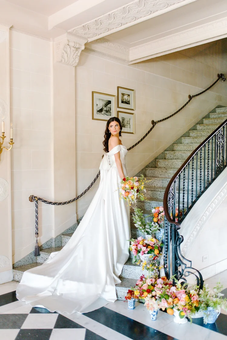 Bride in an off-the-shoulder satin A-line wedding gown with cathedral train standing on a grand staircase, looking back over her shoulder, surrounded by lush colorful floral arrangements in blue and white vases
