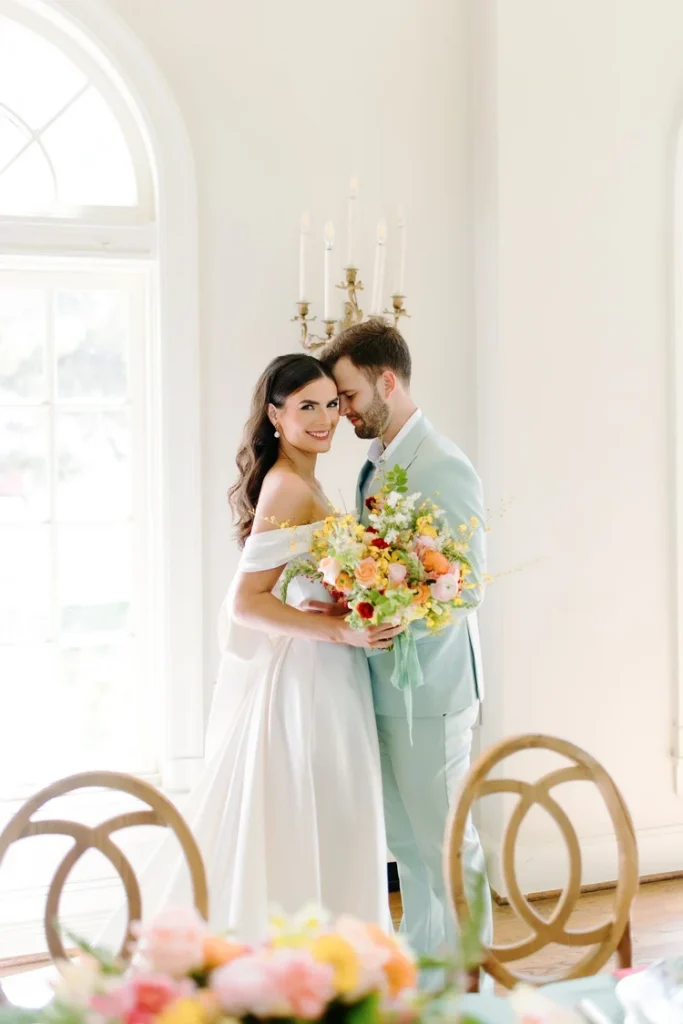 Bride in an off-the-shoulder satin A-line wedding gown holding a colorful bridal bouquet of peach, pink, and yellow florals, embracing her groom in a mint suit inside a bright white event space