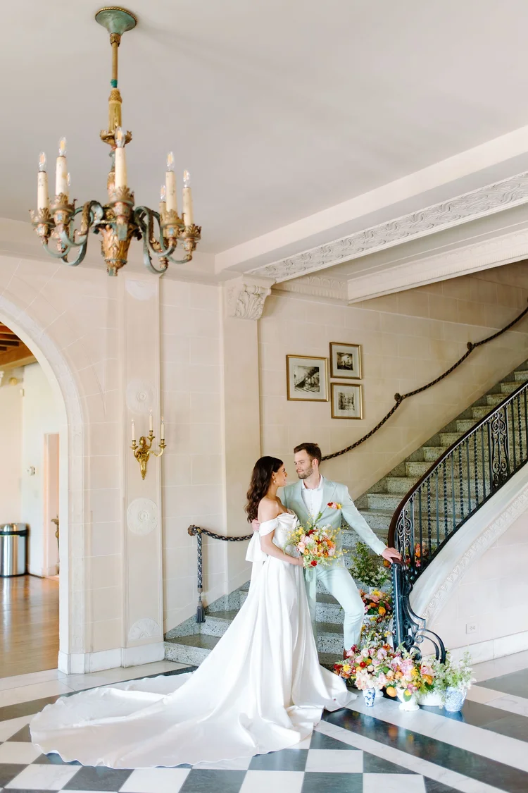 Bride and groom standing together on a black and white checkered marble floor at the base of a grand staircase, bride in an off-the-shoulder satin gown with a cathedral train surrounded by colorful floral arrangements