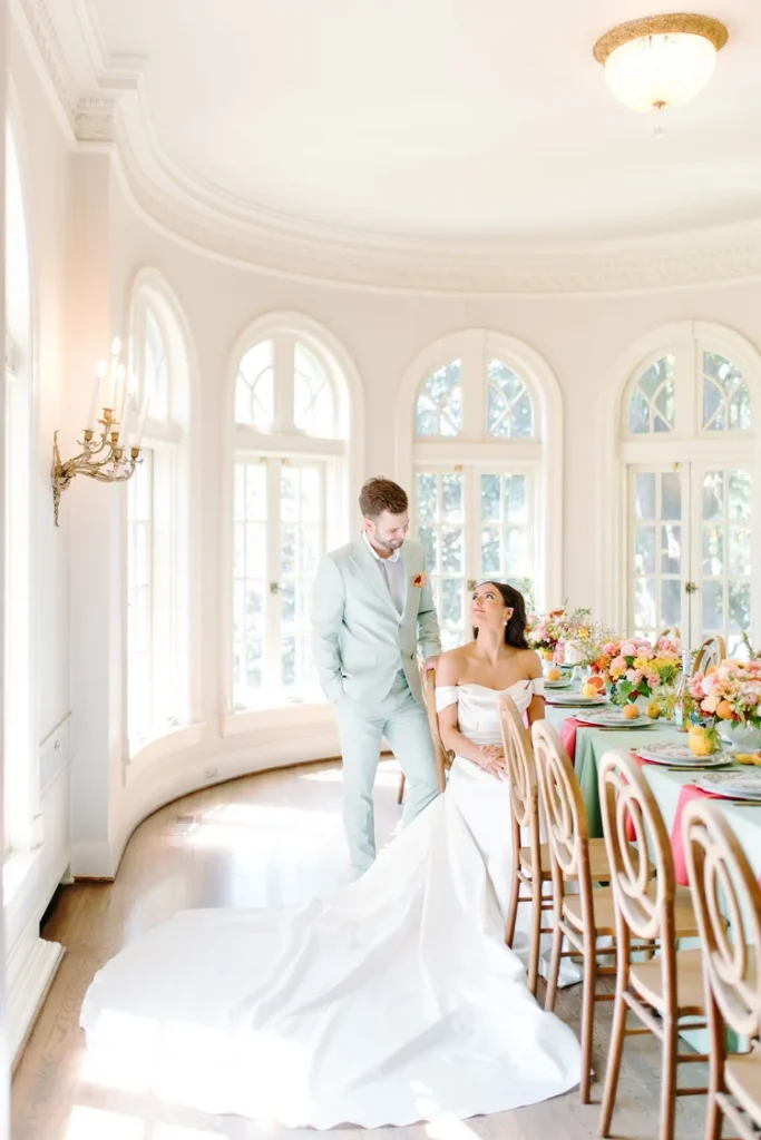 Bride in an off-the-shoulder satin wedding gown with a dramatic cathedral train seated at a colorful reception table, groom in a mint suit standing beside her in a sunlit domed room with arched windows