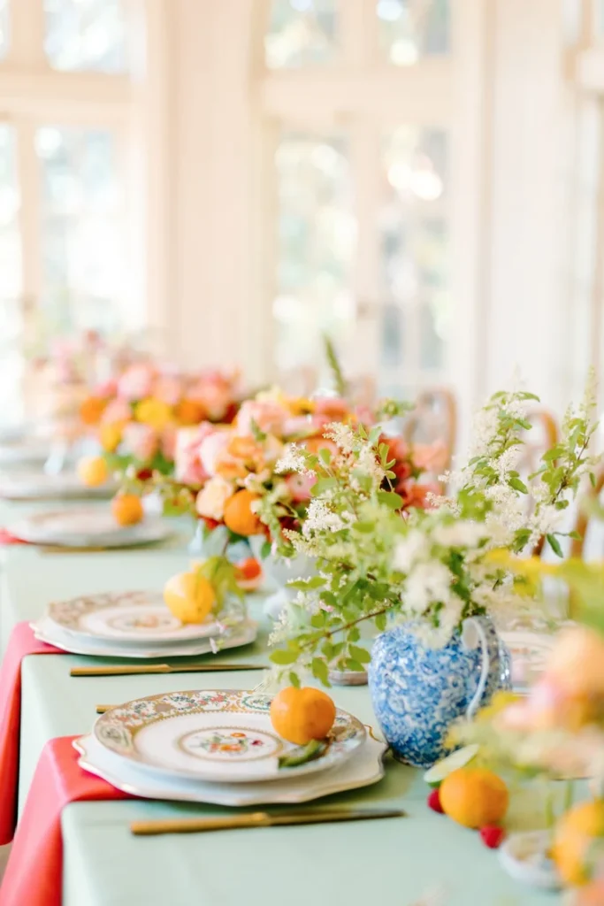Colorful wedding reception tablescape with a sage green linen runner, coral napkins, vintage floral china, blue and white ginger jar vases, and citrus fruit centerpieces with pink and orange blooms