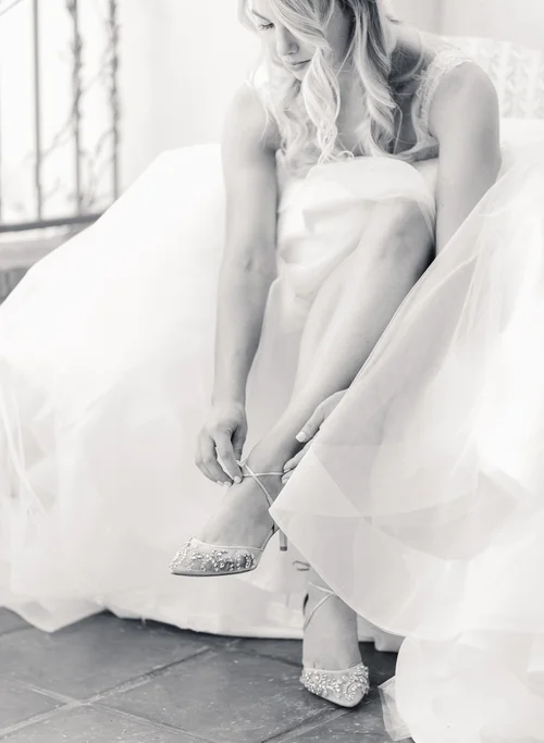 Black and white photo of bride in a tulle ballgown fastening the ankle strap of her Bella Belle beaded bridal heels while getting ready on her wedding day