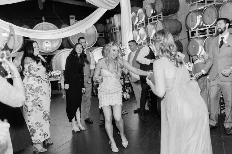Black and white photo of bride dancing joyfully on the reception dance floor in a short beaded dress and Bella Belle shoes, guests cheering around her in a winery venue