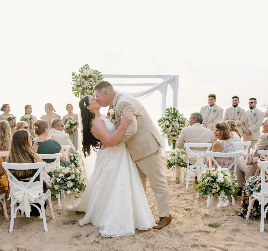 Bride and groom share their first kiss at a beach ceremony in Cozumel, Mexico, surrounded by white chairs, white floral arrangements with monstera leaves, and smiling guests