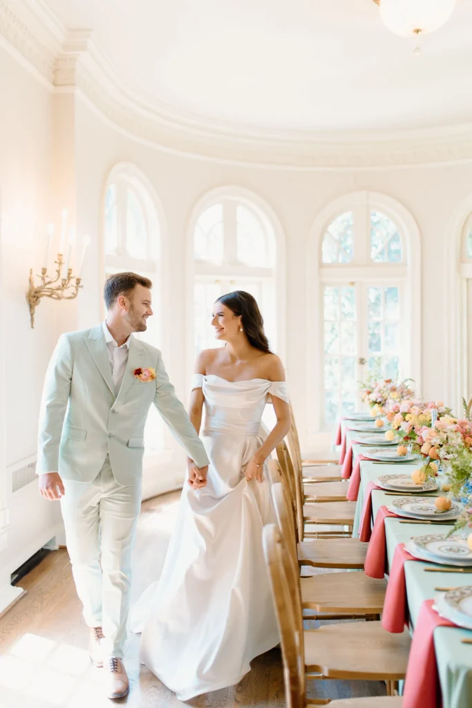 Bride and groom walking hand in hand laughing alongside a colorful wedding reception table in a sunlit white ballroom, bride wearing an off-the-shoulder satin gown from La Mariée Boutique in Tulsa